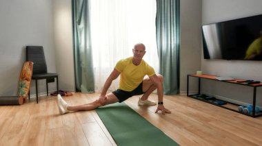 Tam boy kel adam, erkek koç kameraya bakıyor, gündüz evde oturma odasında antrenman yaparken bacaklarını esnetiyor.