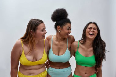 Farklı vücut şekillerine sahip üç farklı genç kadın renkli iç çamaşırı giyiyorlar. Neşeli görünüyorlar, arka planda birlikte poz veriyorlar.