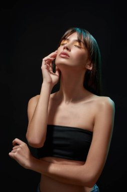 Genç esmer kadın model, renkli makyajlı, sutyen takıyor, başını kaldırıyor, gözleri kapalı poz veriyor, siyah arka plan üzerine izole edilmiş.