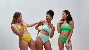 Farklı vücut şekillerine sahip güzel genç kadınlar renkli iç çamaşırları giyiyorlar duygusal görünüyorlar, stüdyoda beyaz arka planda birlikte poz verirken eğleniyorlar.