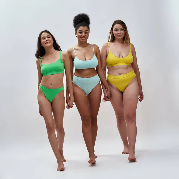 Full length shot of three diverse women in colorful underwear holding hands, looking at camera, standing together isolated over light background