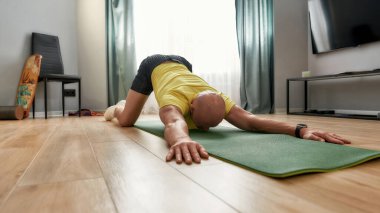 Evde yoga yapan, çocuk egzersizi yapan, Balasana 'da dinlenen, oturma odasında hasırla çalışan kel bir adam.
