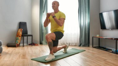 Formunu koru. Evde spor kıyafetleriyle egzersiz yapan olgun bir adam. Kafkas fitness eğitmeni, üniformalı antrenör oturma odasındaki yoga minderine çömelmiş.