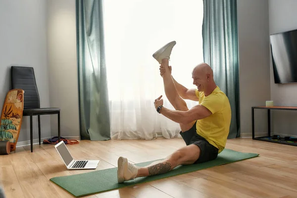 Virtual fitness class. Mature personal fitness conducting online training while sitting on yoga mat at home in the living room, looking at laptop and smiling