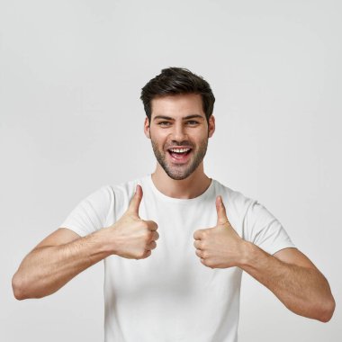 Tişört giyen sakallı adam başparmaklarını gösteriyor.