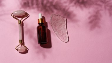 Gül kuvars sha taşı, yeşim silindir ve cam göz damlası serumu şişesi pembe yüzeyde çapraz izole bir şekilde duruyor.