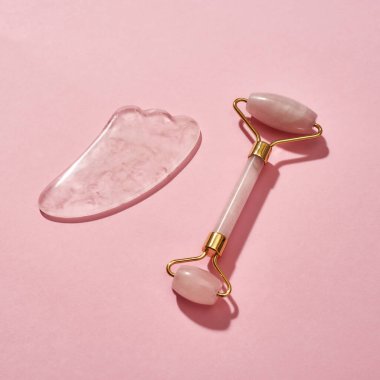 Gül kuvars yüz masajı aletleri, yüz silindiri ve pembe arka planda izole edilmiş Gua Sha kazıyıcısı.