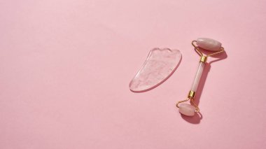 Gül kuvars yüz masajı aletleri, yüz silindiri ve pembe arka planda izole edilmiş Gua Sha kazıyıcısı.