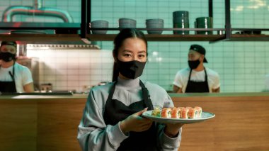 Koruyucu maske takan genç bir garson kameraya bakıyor ve elinde suşi ruloları olan bir tabak tutuyor. Restoranda Japon mutfağı..