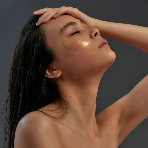 Νεαρή όμορφη Ασιάτισσα ημίγυμνη γυναίκα σε πετσέτα μπάνιου με καλλυντικά μπαλώματα ματιών αγγίζοντας το πρόσωπό της, ενώ τα μάτια κλειστά — Φωτογραφία Αρχείου