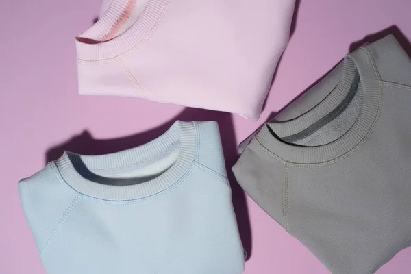 Closeup shot of three folded pastel pink, blue and gray sweatshirts isolated on pale purple background — Stock Photo, Image