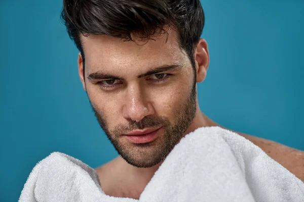 Studio portrét sexy brunetky muž s mokrým umytým obličejem drží bílý ručník, při pohledu na kameru, pózování izolované přes modré pozadí — Stock fotografie