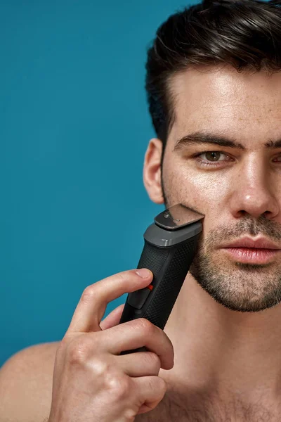 Half face closeup of brunette handsome guy looking at camera while using electric shaver isolated over blue background — Stock Photo, Image