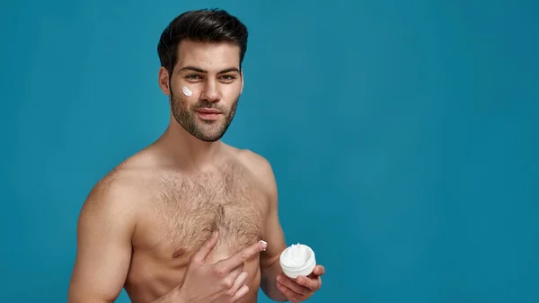 Studio shot van sexy brunette man reclame, wijzend op witte pot effectieve crème in zijn hand, kijken naar de camera, poseren geïsoleerd over blauwe achtergrond — Stockfoto