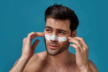 Kahverengi saçlı, beyaz maskeli, gözenekleri temizlemek için burnuna sürtünmüş, mavi arka planda yalnız poz veren kendine güvenen bir adam.