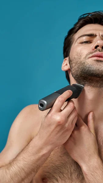 Портрет брюнетки голый мужчина выглядит сфокусированным при использовании электрической бритвы изолированы на синем фоне — стоковое фото