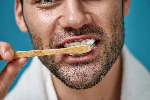 Cropped shot of handsome man with towel around his neck brushing his teeth using wooden toothbrush isolated over blue background