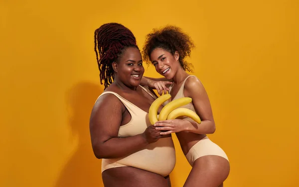 Due giovani donne africane americane carine in biancheria intima sorridenti alla macchina fotografica, con in mano banane gialle mature, in posa insieme isolate su sfondo arancione — Foto Stock