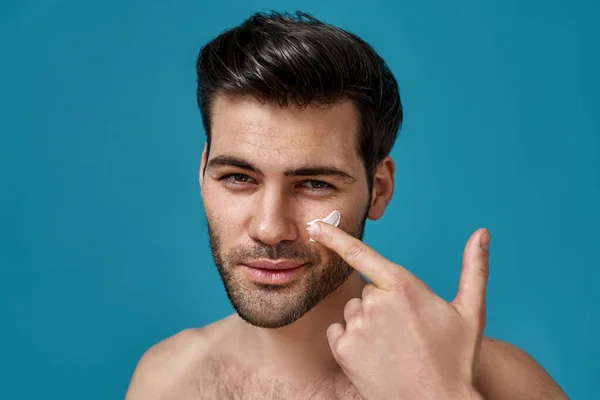 Close-up portret van goed uitziende naakt brunette guy toepassen effectieve crème op zijn gezicht huid terwijl kijken naar camera, poseren geïsoleerd over blauwe achtergrond — Stockfoto
