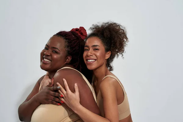 Ritratto di due donne africane americane sorridenti in biancheria intima con diverse dimensioni del corpo che si divertono mentre posano insieme isolate su sfondo grigio — Foto Stock