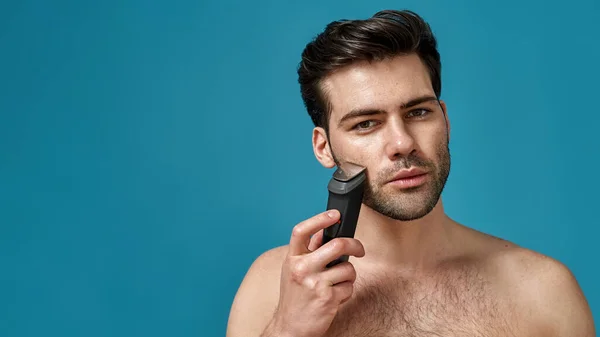Beauty portrait of handsome young man using electric shaver isolated over blue background — Stock Photo, Image