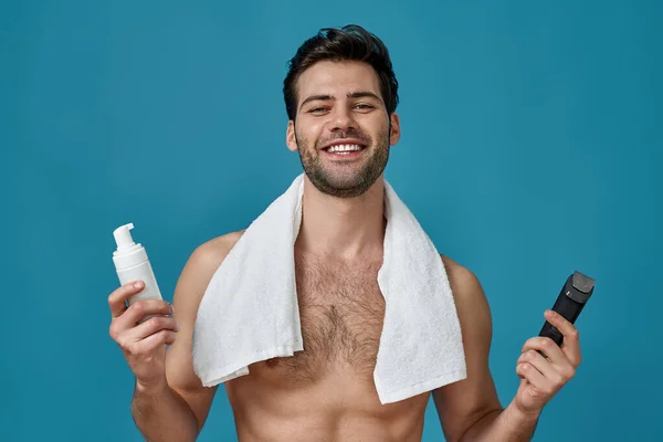 Aantrekkelijke naakte man met witte handdoek om zijn nek glimlachend op de camera, met een scheerschuim en elektrisch scheerapparaat, poseren geïsoleerd over blauwe achtergrond — Stockfoto