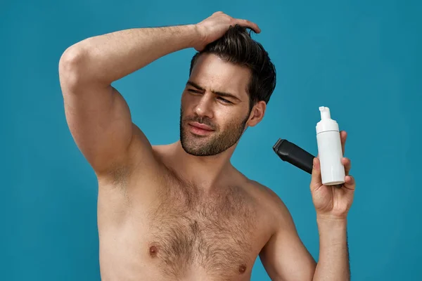 Handsome naked guy with hand on his hair looking away, holding a shaving foam and electric shaver isolated over blue background — Stock Photo, Image