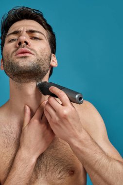 Mavi arka planda izole edilmiş elektrikli traş makinesi kullanırken konsantre olmuş görünen yakışıklı adamın portresi.