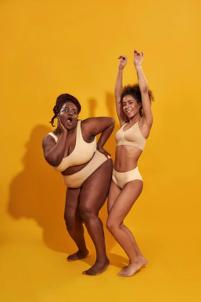 In voller Länge Aufnahme fröhlicher afrikanisch-amerikanischer Frauen in beiger Unterwäsche mit unterschiedlicher Körpergröße, die verspielt aussehen, während sie isoliert auf gelbem Hintergrund posieren — Stockfoto