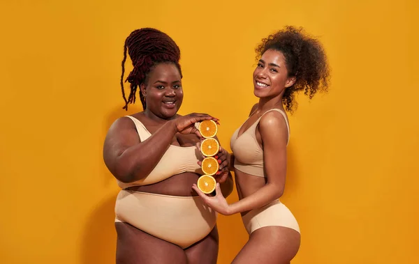 Studio shot di allegre donne africane americane in biancheria intima sorridente mentre tengono mature metà arancione succosa, posa isolata su sfondo arancione — Foto Stock