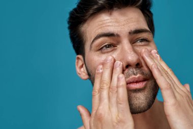 Yüzüne nemlendirici krem sürerken yana bakan yakışıklı bir adamın mavi arka planda izole edilmiş portresi.