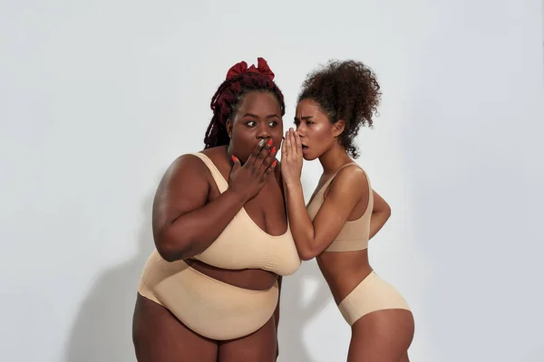 Afro-Amerikaanse vrouw in ondergoed delen van een geheim met haar plus size vriend, staan samen geïsoleerd over grijze achtergrond — Stockfoto