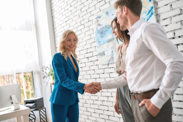 Çekici kadın emlakçı, modern ofiste dururken genç bir çiftle el sıkışıyor.