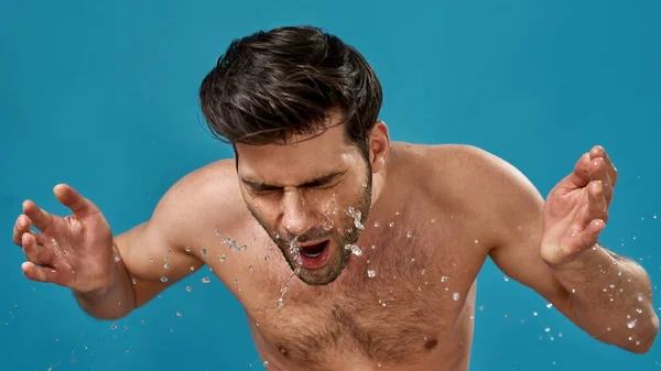 Retrato de un joven guapo lavándose la cara con muchas salpicaduras de agua alrededor aislado sobre fondo azul —  Fotos de Stock