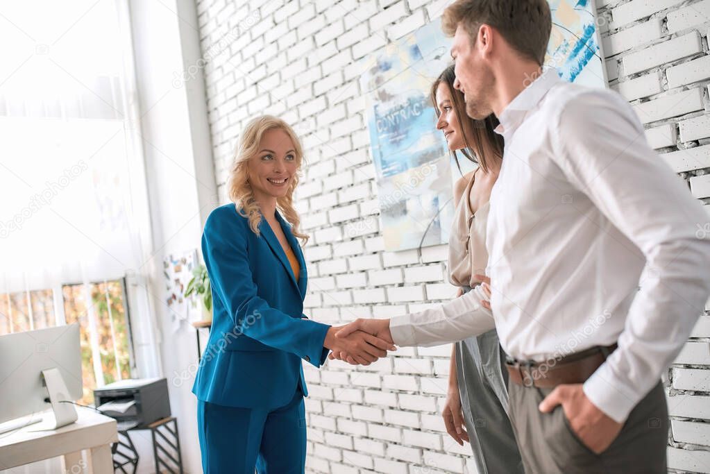 Attractive female realtor shaking hands, making a deal with young couple while standing in modern office