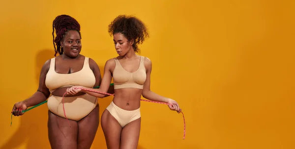 Studio shot van twee Afro-Amerikaanse vrouwen in ondergoed met verschillende lichaamsvorm met metrische meetlint, geïsoleerd over oranje achtergrond — Stockfoto