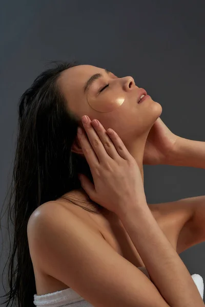 Jonge mooie aziatische half naakte vrouw in bad handdoek met cosmetische ooglapjes op het gezicht aanraken van haar gezicht en het houden van ogen gesloten terwijl poseren tegen donkere achtergrond — Stockfoto