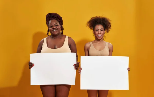 Dos alegres mujeres afroamericanas diferentes en ropa interior riéndose de la cámara, sosteniendo banner en blanco para su texto, de pie aislado sobre el fondo amarillo — Foto de Stock