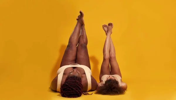 Δύο νεαρά αφροαμερικανικά θηλυκά μοντέλα με εσώρουχα ξαπλωμένα στο πάτωμα με τα πόδια τους πάνω απομονωμένα σε κίτρινο φόντο. — Φωτογραφία Αρχείου