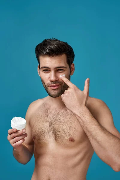 Sexy naked brunette guy applying effective cream in white jar on his face while looking at camera, posing isolated over blue background — Stock Photo, Image