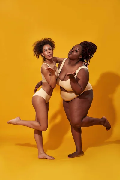 Cropped shot of two confident african american female models in underwear  with different body weight holding hands on waist, standing together  isolated over yellow background Stock Photo by ©LanaStock 481035630
