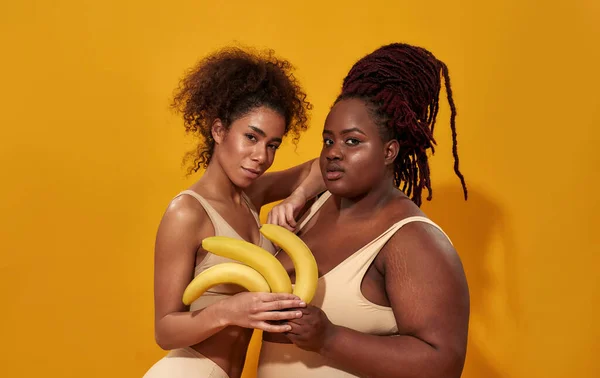 Due giovani donne africane americane in biancheria intima guardando la macchina fotografica, tenendo in mano banane gialle mature, in posa insieme isolate su sfondo arancione — Foto Stock
