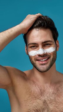 Beyaz maskeli, gülümseyen esmer adam, kahverengi saçlarına dokunan gözenekleri temizlemek için burnuna sürtünmüş, mavi arka planda izole bir şekilde poz vermiş.