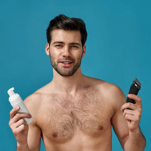 Beauty portrait of sexy brunette guy looking at camera, holding electric shaver and shaving foam for better shave and to protect skin from irritation isolated over blue background — Stock Photo, Image