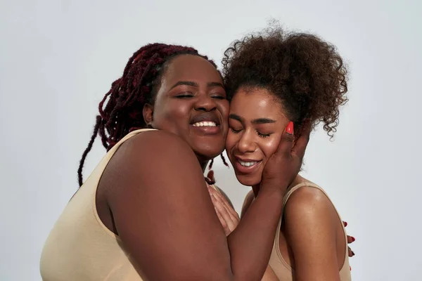 Ritratto di amici felici, donne afro-americane in biancheria intima con diverse dimensioni del corpo sorridenti, che si abbracciano mentre posano insieme isolati su sfondo grigio — Foto Stock