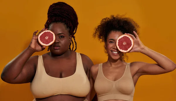 Studio porträtt av två unga afrikanska amerikanska kvinnor i underkläder tittar på kameran, håller hälften av saftig grapefrukt över ett öga, poserar isolerad över orange bakgrund — Stockfoto