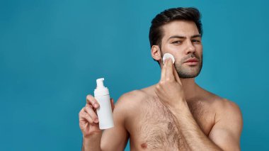 Kendine güvenen esmer, sabah rutini yapan, pamuk pedlerle yüzünü temizleyen, mavi arka planda izole edilmiş köpük temizleyicisi reklamcısı.