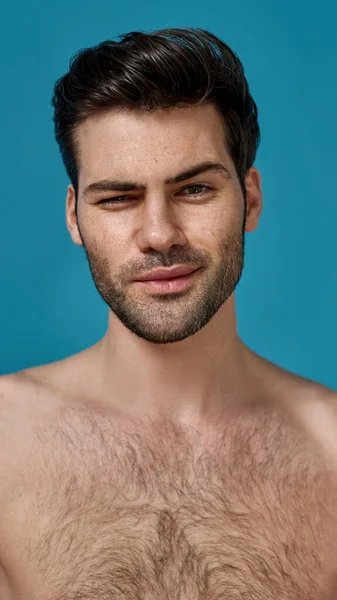Porträt eines attraktiven nackten, brünetten Mannes mit frischer, perfekt glatter Haut vermasselt ein Auge, während er die Kamera isoliert vor blauem Hintergrund betrachtet — Stockfoto