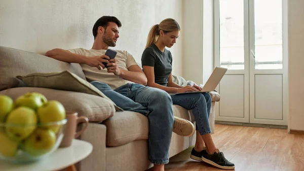 Pareja joven sentada en el sofá y tratando de completar la tarea de las mujeres —  Fotos de Stock