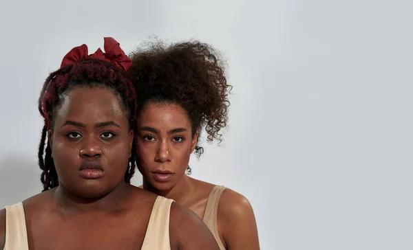 Closeup portrait of two serious african american women with different body size looking at camera, posing together isolated over gray background — Stock Photo, Image